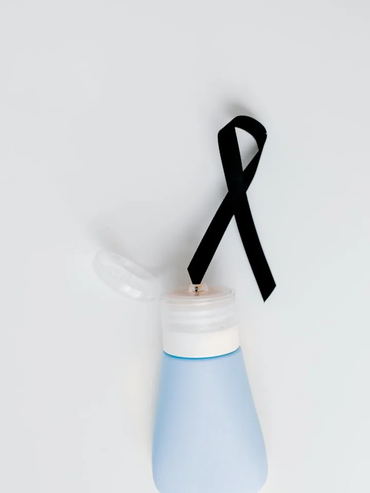 Light blue sunscreen bottle with an open cap and a black awareness ribbon draped behind it on a light, neutral background.