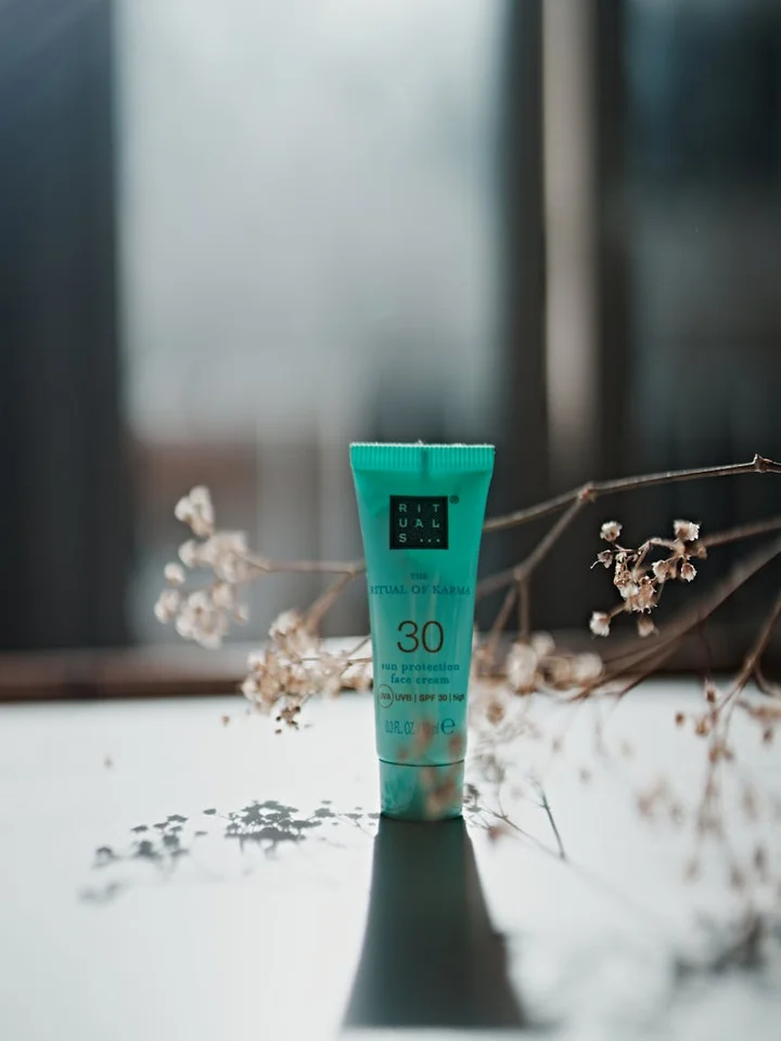 Turquoise SPF 30 sunscreen tube standing on a reflective surface with a blurred window background and dried branches nearby.