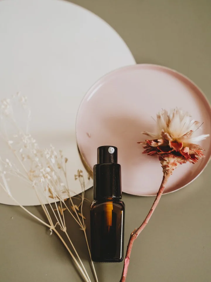 Amber glass bottle with a spray top placed beside a soft pink plate and dried botanical stems on a neutral background, illustrating the mix of carrier oils and botanical extracts in skincare.