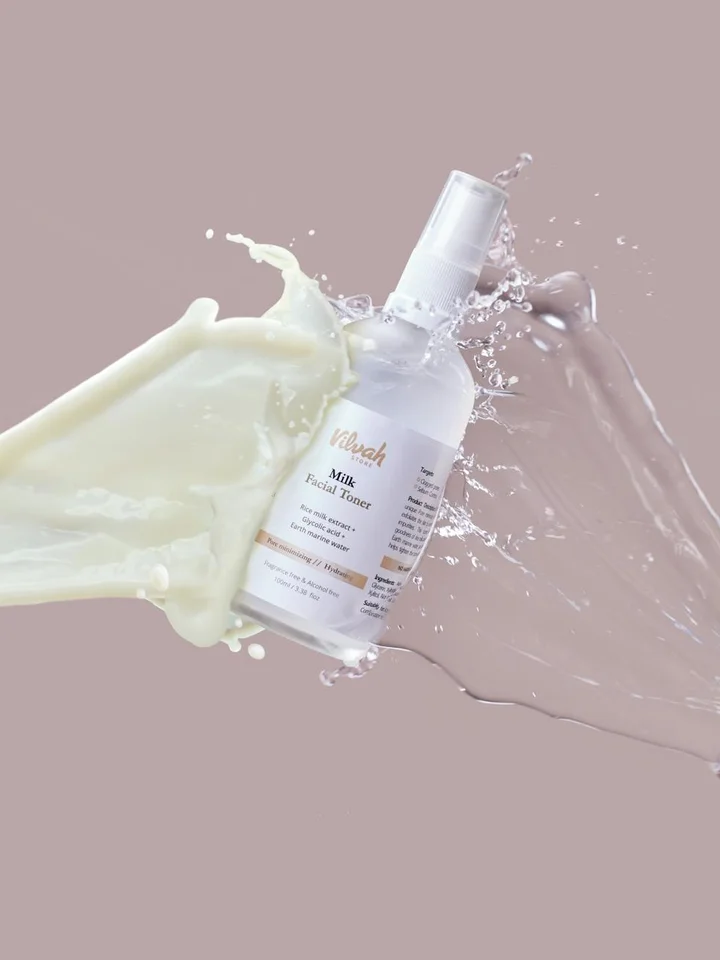 Bottle of toner being sprayed with liquid splashes on a soft pink background