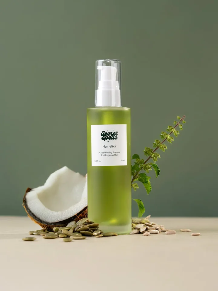 A translucent green bottle with a pump dispenser sits on a light beige surface beside a halved coconut, scattered seeds, and a sprig of herb against a muted green background.