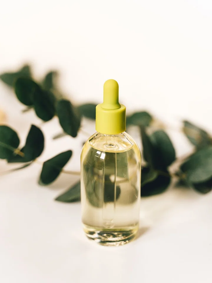 Dropper bottle with pale yellow serum on a white surface, green leaves blurred in the background.