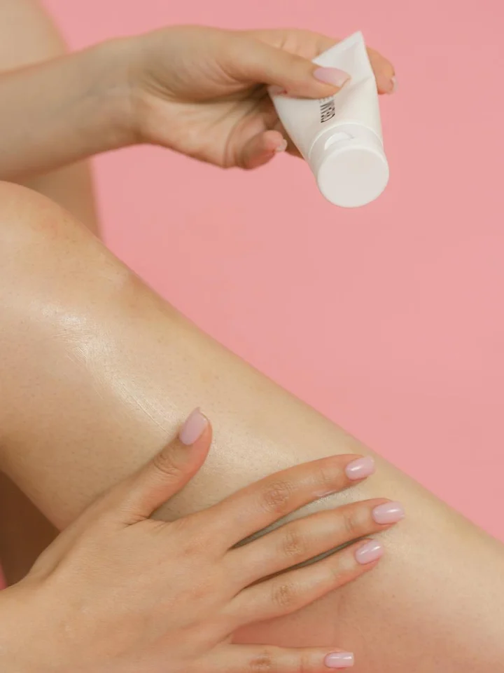 Close-up of a person applying a pea-sized amount of cream to the leg against a pink background