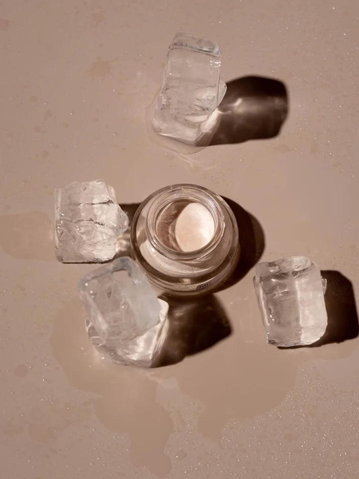 Open glass jar of cream on a beige surface, surrounded by several ice cubes