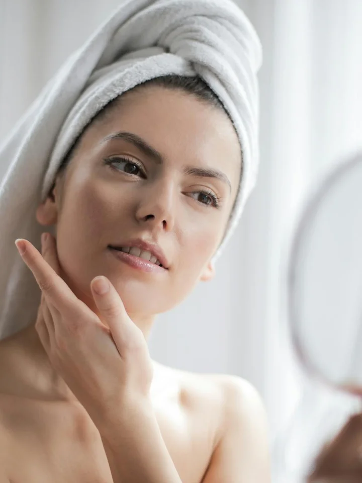 Woman with a towel wrapped around her head, looking into a mirror and touching her cheek during a skincare routine.