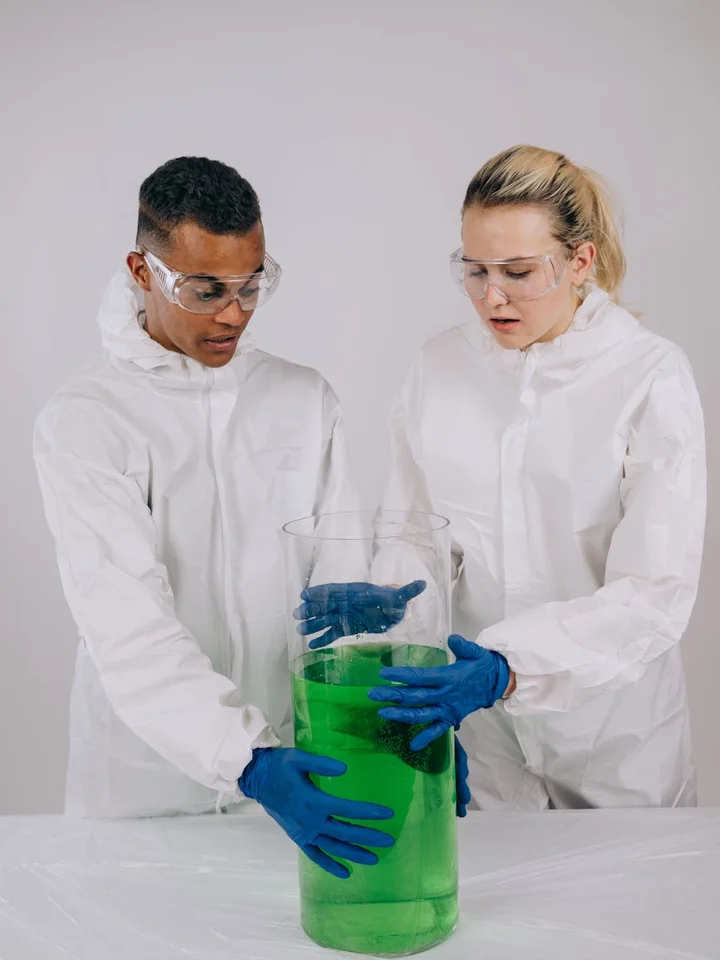 Two scientists in white lab coats and safety goggles inspect a large beaker filled with bright green liquid in a laboratory.
