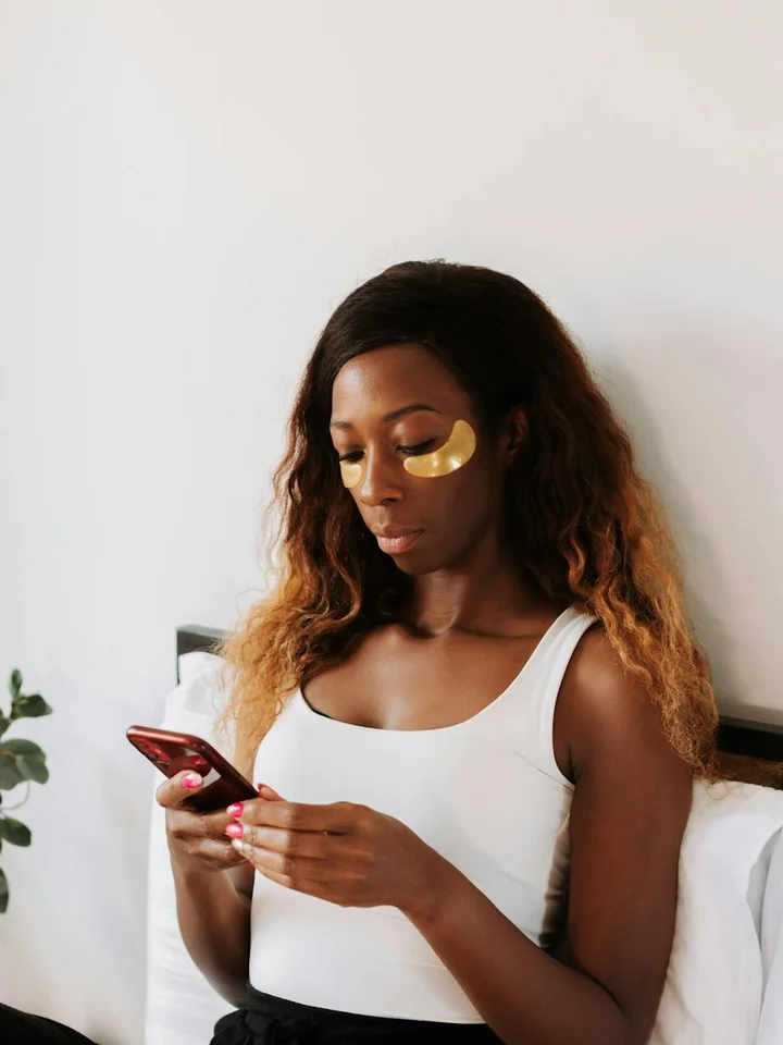 A woman with long wavy hair wearing a white tank top sits against a light background, applying gold under-eye patches while looking at her phone.