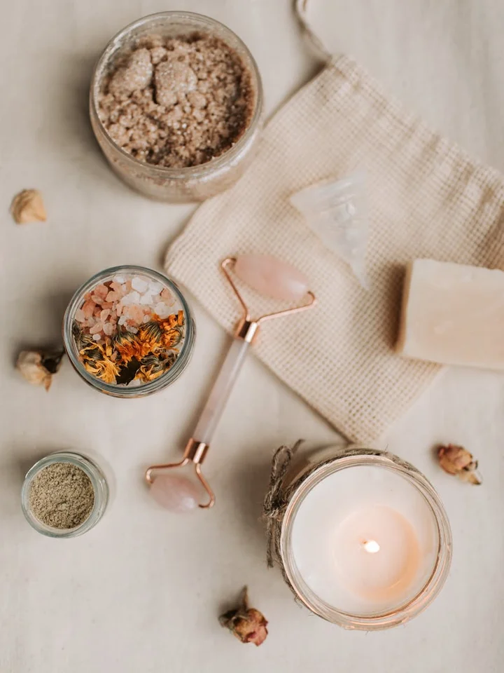 Flat lay of skincare items on a light surface: a candle in a glass jar, a rose-quartz facial roller, a bar of soap, and small jars containing bath salts and dried botanicals.