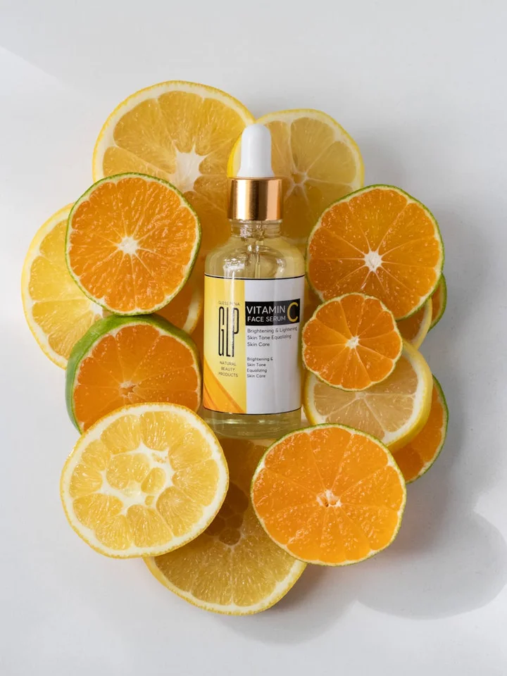 A small glass dropper bottle labeled Vitamin C sits on a white surface, surrounded by orange and lemon halves.