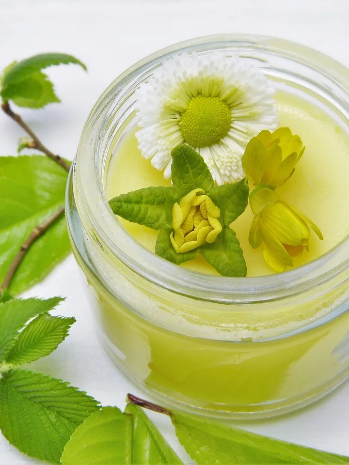 Close-up of a pale yellow moisturizer in a glass jar with white and yellow flowers and green leaves surrounding it