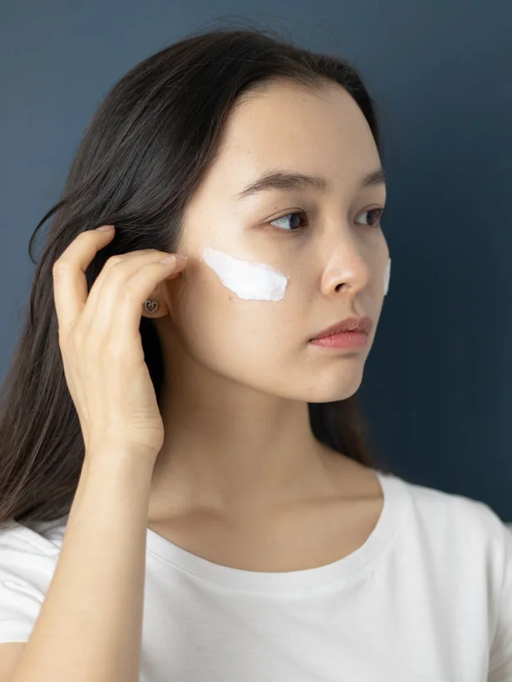 A young woman applies a thin layer of cream to her cheek with her fingers, preparing to use a topical treatment.