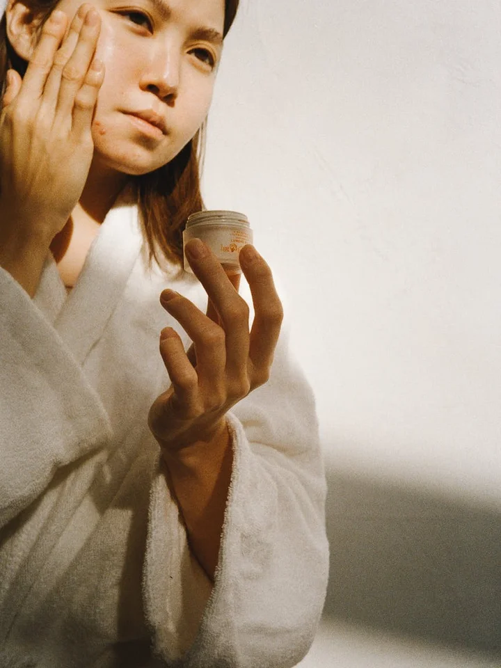 A person in a white robe applies cream to their face, holding a small jar in their hand.