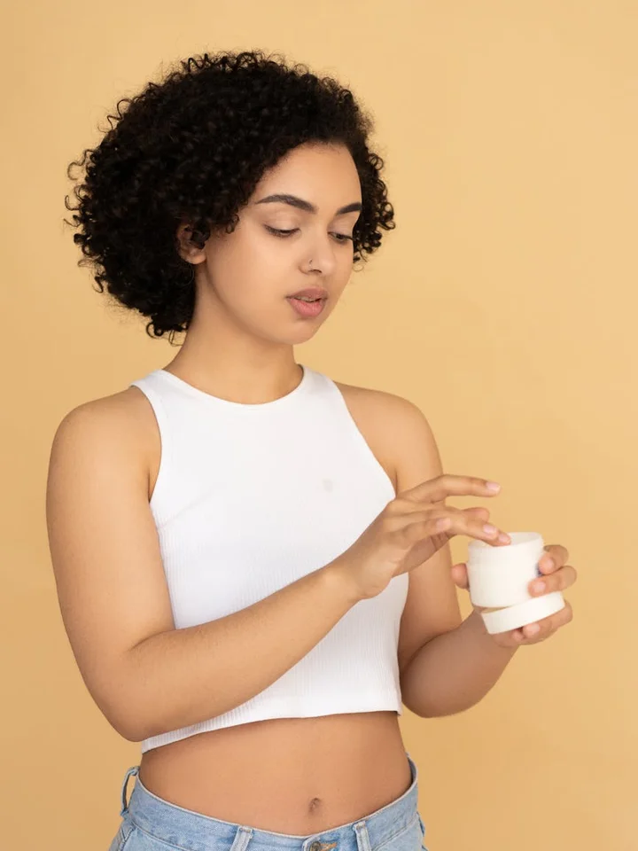 A woman wearing a white sleeveless crop top applies moisturizer from a small jar, preparing her skin for self-tanning.