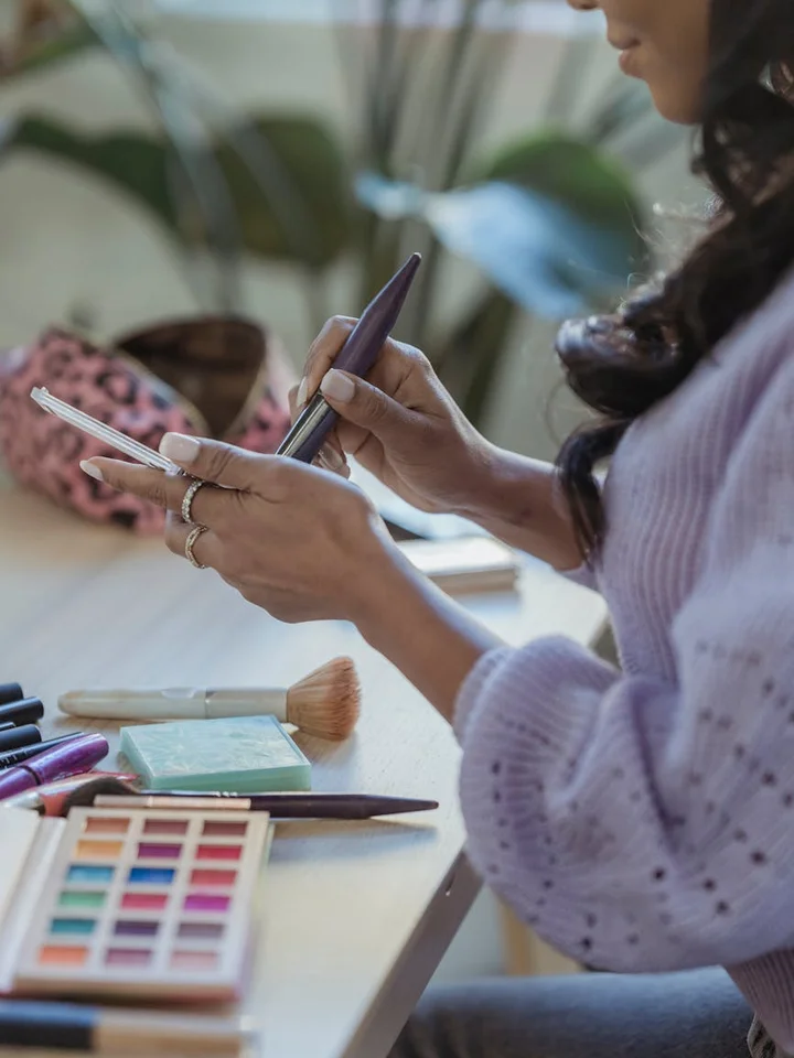 A person sits at a vanity, holding a makeup brush in one hand and a smartphone in the other, with an eyeshadow palette and brushes on the table.