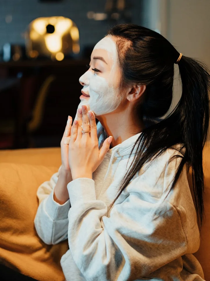 Woman applying a white facial mask at home while seated on a sofa, wearing a light gray hoodie.