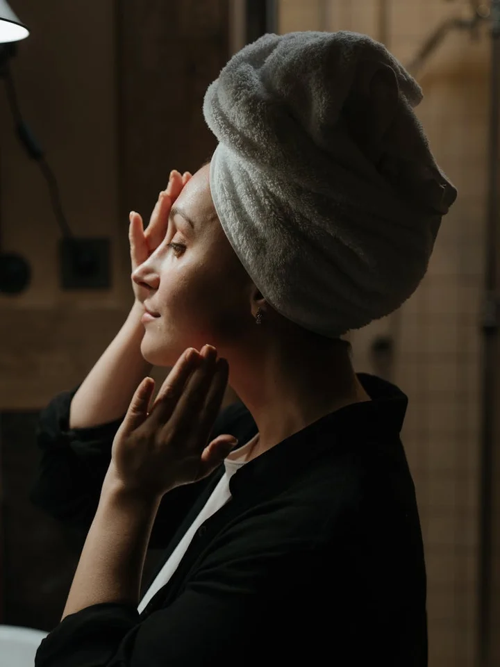 Side profile of a person with a towel wrapped around their head in a bathroom, preparing a skincare routine.