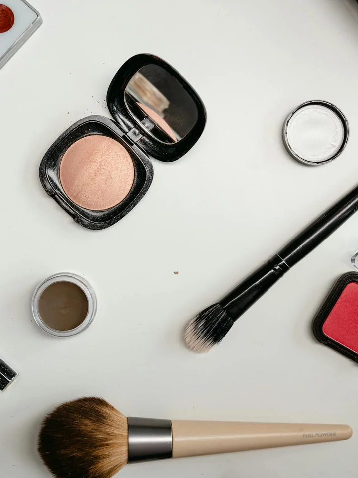 Flat lay of makeup essentials on a white surface: compact powder, blush, and makeup brushes, illustrating foundation products and tools related to acne-prone skin.